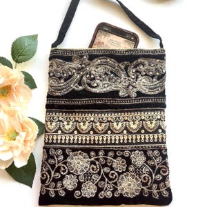 May include: Black velvet crossbody bag with ornate silver embroidery and sequin details. The bag features a long black strap and a phone is visible inside. Floral and scrollwork patterns adorn the bag's exterior.