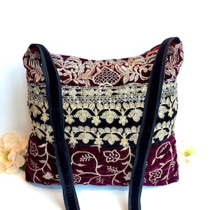 May include: A burgundy velvet shoulder bag with gold embroidery and black velvet straps. The bag has a floral design and a black sequined band. A stylish accessory.