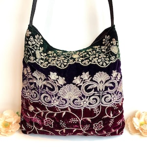 May include: A velvet shoulder bag in shades of green, purple, and burgundy, with gold floral embroidery. The bag features a black strap. The bag's design showcases a gradient of colours.