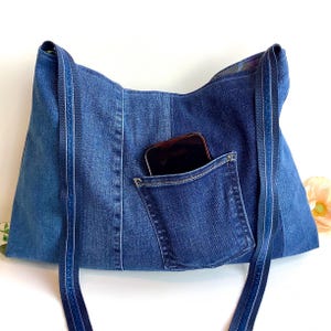 May include: A denim tote bag with a pocket and straps. The bag is made from recycled blue jeans. A dark-colored smartphone is in the pocket. The bag has a light blue and dark blue color scheme.