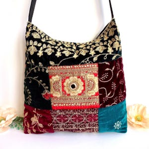 May include: A patchwork shoulder bag with a black strap. The bag features a variety of fabrics in jewel tones, including black velvet with gold embroidery, deep red velvet, and teal velvet. A central red square is adorned with gold embellishments.