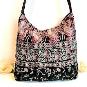 May include: A black shoulder bag with detailed embroidery. The bag showcases a paisley pattern in red and gold, floral designs, and a dark green velvet base. The strap is black.