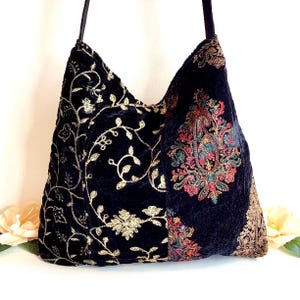 May include: A black velvet hobo bag featuring intricate gold embroidery in a scrolling vine pattern on the left side and a colorful, detailed paisley motif in shades of red, green, and gold on the right side, complete with a black shoulder strap.