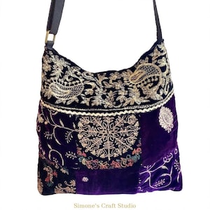 Peut inclure: Un sac à bandoulière en velours violet profond orné de broderies dorées complexes aux motifs floraux et cachemire. Le sac possède une sangle noire et une attache en métal doré. Le texte Simone's Craft Studio est imprimé en bas.