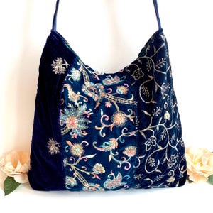 May include: A dark blue velvet hobo bag featuring a central panel with multi-colored floral embroidery and side panels adorned with intricate gold-toned vine and leaf stitching. The bag includes a matching dark blue shoulder strap.