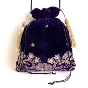 May include: A deep purple velvet drawstring pouch featuring gold metallic floral embroidery along the bottom edge. The bag includes two long black cord straps with gold-toned beads at the ends and two decorative beige tassels on the sides.
