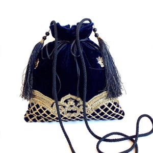 May include: A deep navy blue velvet drawstring pouch featuring intricate gold metallic embroidery in a lattice pattern at the base. The bag includes black braided cord straps, black beaded accents, and long black tassels on each side.