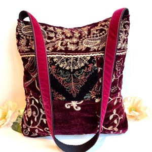 May include: A burgundy velvet tote bag with gold and cream floral embroidery. The bag has a black strap and a deep red velvet shoulder strap. The bag is decorated with intricate embroidery and has a soft, luxurious appearance.