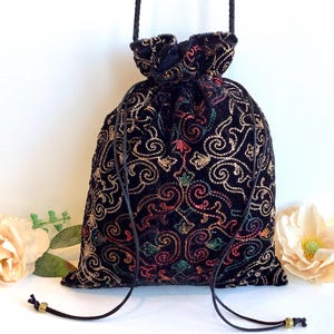 May include: Black velvet drawstring bag with gold, red, and green embroidered floral and swirl design. The bag has a black cord drawstring closure and a black cord handle. The bag is approximately 20 cm tall and 15 cm wide.