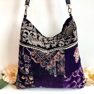 May include: A deep purple velvet hobo bag featuring intricate gold and multi-coloured floral embroidery in a patchwork design. The bag includes a black fabric shoulder strap attached with antique brass-toned metal swivel clasps and rings.