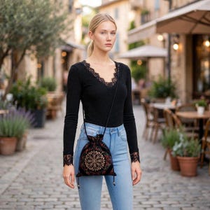 May include: A black long-sleeved ribbed top with a V-neckline featuring lace trim and lace cuffs, paired with light blue denim jeans and a small black drawstring pouch bag decorated with a gold-colored circular embroidered mandala pattern.