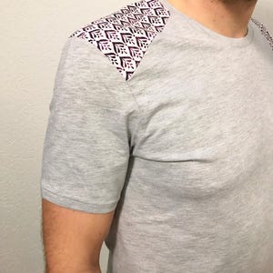 Può includere: Una maglietta grigia a maniche corte con un dettaglio a fantasia sulla spalla. Il dettaglio sulla spalla presenta un motivo ripetuto in bianco e viola scuro. La maglietta è realizzata in un materiale morbido.