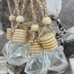 May include: A collection of small, clear glass bottles with geometric, diamond-like facets. Each bottle features a light-colored, ribbed wooden cap and is suspended by a braided beige cord interwoven with thin, shimmering gold metallic threads.