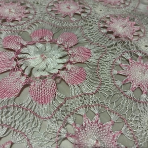 May include: Close-up of a white and pink crocheted lace fabric. The design features floral patterns with pink accents and a delicate, open weave. The lace is likely used for decorative purposes.
