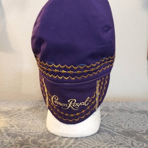 May include: A purple hat with gold embroidered trim and the words "Crown Royal" in gold script. The hat is displayed on a white mannequin head. The hat has a curved brim and a rounded crown.