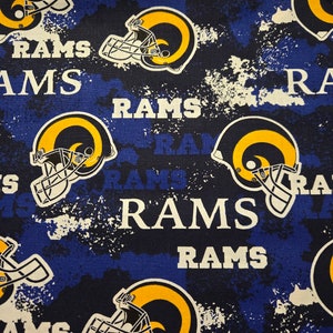 May include: Fabric with a repeating pattern of Los Angeles Rams football helmets in yellow, white, and navy blue. The word "RAMS" is printed in white across the navy blue background, with a distressed, splattered effect.