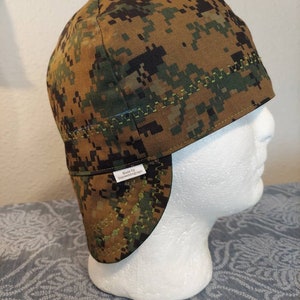 May include: A camouflage welding cap in shades of brown, green, and black, with a digital pixel pattern. The cap has a curved brim and is stitched with a contrasting green thread. The tag reads "Made by Dryweldingcaps".