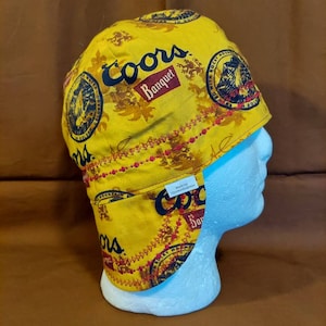 May include: Yellow and brown welding cap with the Coors Banquet logo and other graphics. The cap is on a white mannequin head. The cap has a red stitched trim and the words "Made by Tinywendyspace" on a white tag.