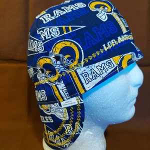 May include: A blue and yellow Los Angeles Rams welding cap. The cap features the team's logo, helmet graphics, and the word "RAMS" in white. The cap has a brim and ear flaps. The text "Made by Diyweldinngage" is visible.