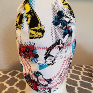 May include: White fabric cap featuring colorful robot illustrations in yellow, blue, and red. The cap has a red stitched trim and is displayed on a white mannequin head. The design includes comic book style elements.