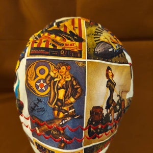 May include: A white surgical cap with a patchwork design featuring vintage aviation pin-up art. The cap has a red embroidered trim and panels with images of airplanes and women in retro attire. The text "REPUBLIC AVIATION" is visible.