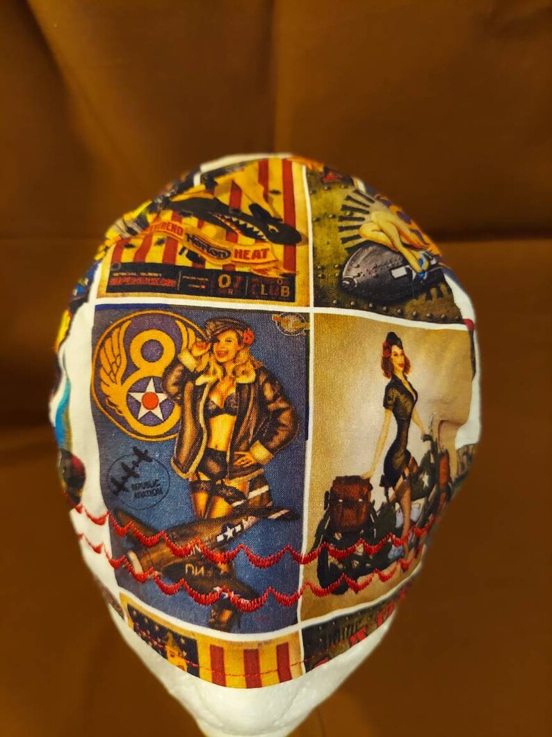 May include: A white surgical cap with a patchwork design featuring vintage aviation pin-up art. The cap has a red embroidered trim and panels with images of airplanes and women in retro attire. The text "REPUBLIC AVIATION" is visible.