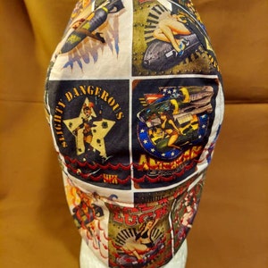 May include: A white and black patterned cap featuring vintage-style pin-up girl and military aircraft graphics. The cap has a patchwork design with the words "Slightly Dangerous" and "American Luck" on it. The cap is displayed on a white foam head.