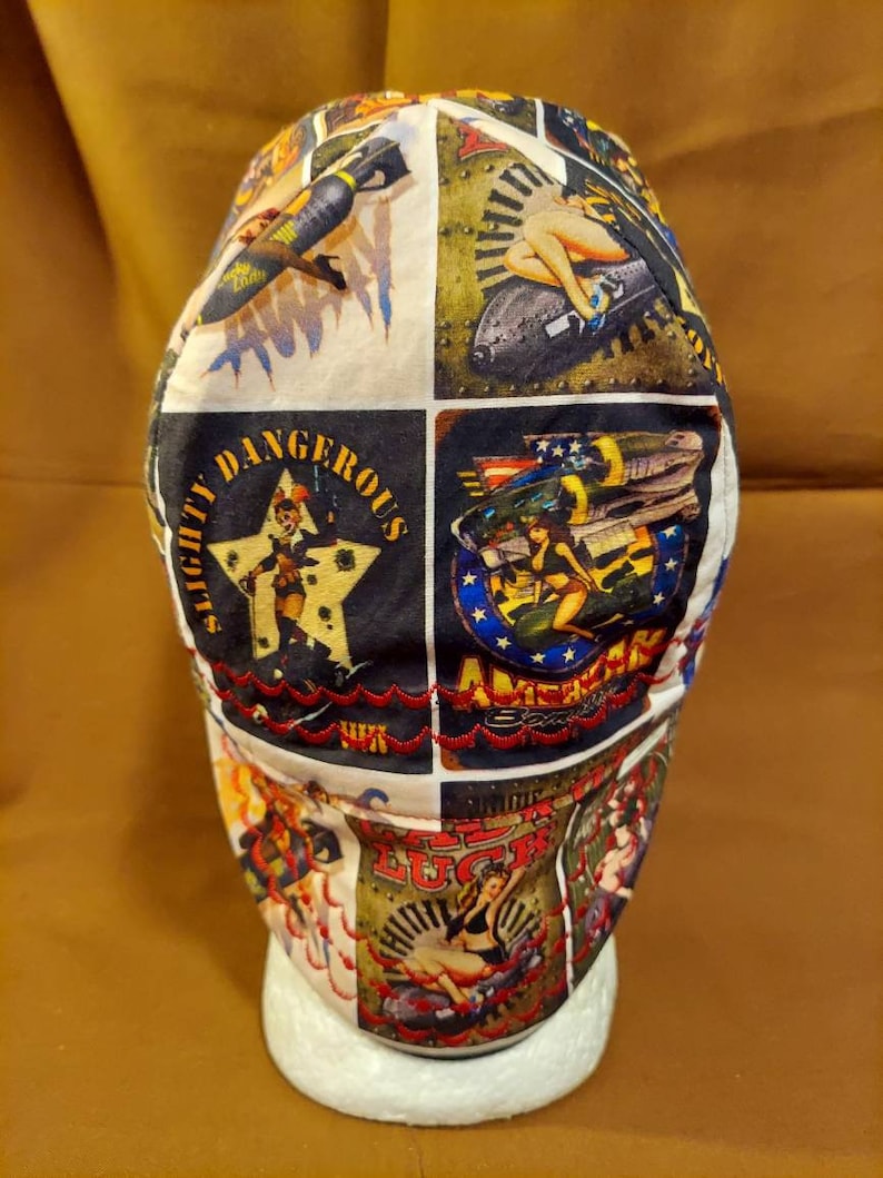 May include: A white and black patterned cap featuring vintage-style pin-up girl and military aircraft graphics. The cap has a patchwork design with the words "Slightly Dangerous" and "American Luck" on it. The cap is displayed on a white foam head.