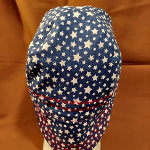 May include: A blue and white star-patterned cap with a red stitched brim. The cap is made of fabric and features a repeating pattern of white stars on a navy blue background. The brim is decorated with red stitching.