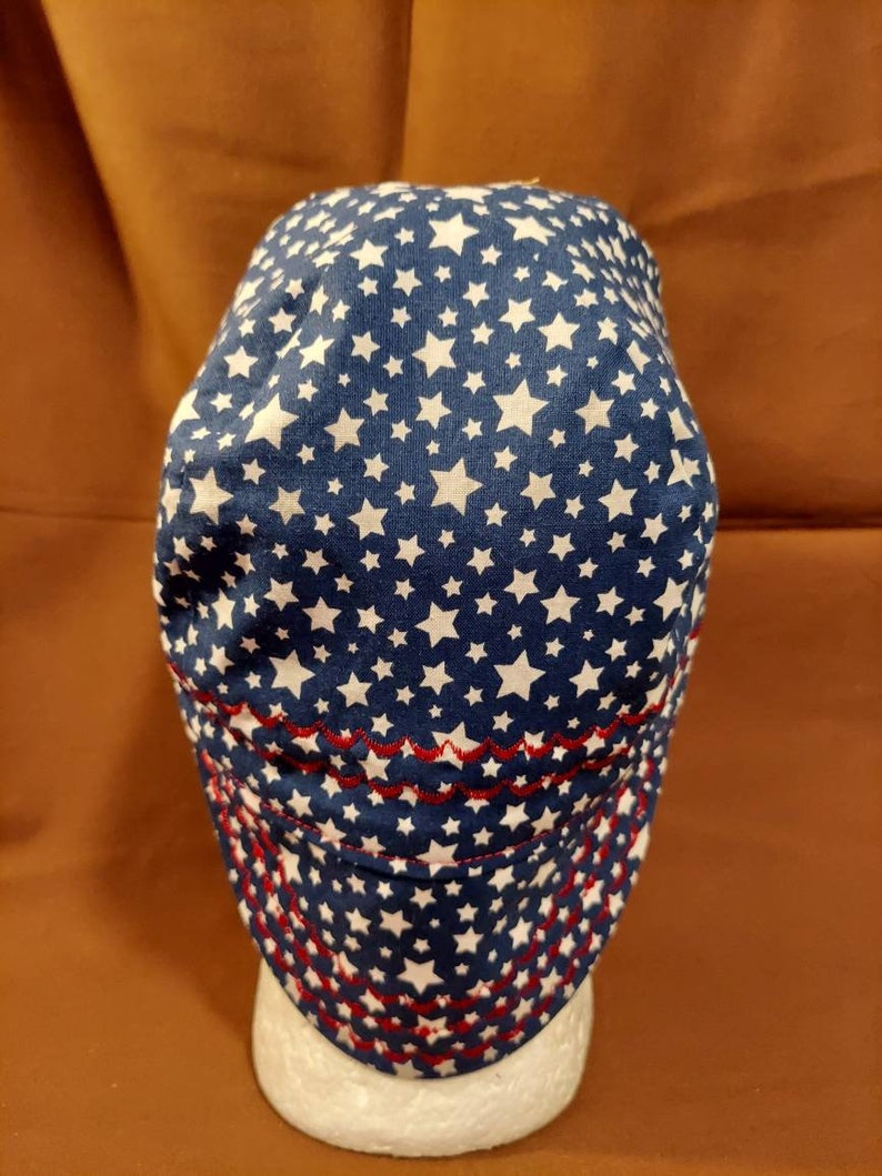 May include: A blue and white star-patterned cap with a red stitched brim. The cap is made of fabric and features a repeating pattern of white stars on a navy blue background. The brim is decorated with red stitching.