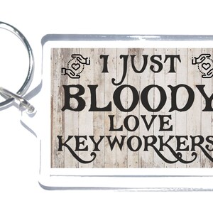 Könnte beinhalten: Schlüsselanhänger mit weißem Holzmaserungshintergrund und dem Text "I just bloody love keyworkers!" in Schwarz. Zwei Hände mit Herzen befinden sich auf beiden Seiten des Textes.