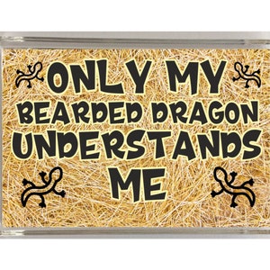 Peut inclure: Un aimant rectangulaire avec un fond de paille et du texte noir qui dit "Only my bearded dragon understands me". Deux silhouettes de lézard noires sont de chaque côté du texte.