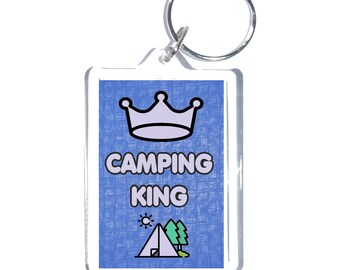 May include: A rectangular keychain with a blue background and the words "CAMPING KING" in white. The keychain features a crown, a tent, trees, and a sun graphic. A silver key ring is attached.