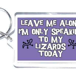 Puede incluir: Un llavero de acrílico morado con un borde blanco y un anillo de llave plateado. El llavero presenta un diseño de texto blanco que dice "Leave me alone I'm only speaking to my lizards today" con dos siluetas de lagarto negras a cada lado del texto.