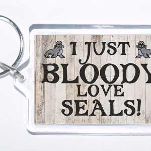 May include: A white wooden background with a black and white graphic that says "I just bloody love seals!" with a cartoon seal on each side of the text. The graphic is on a clear acrylic keychain.