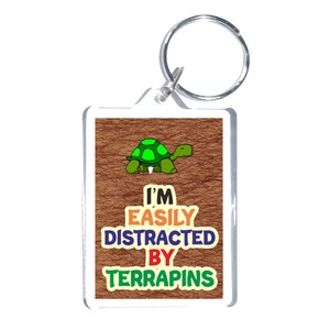 May include: A white acrylic keychain with a brown background and a cartoon turtle. The text on the keychain reads "I'm Easily Distracted By Terrapins".