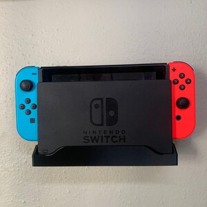 Puede incluir: Una base de Nintendo Switch negra con un Joy-Con azul a la izquierda y un Joy-Con rojo a la derecha. El logotipo de Nintendo Switch es visible en la parte frontal de la base. La base está montada en una pared blanca.