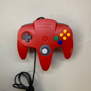 Peut inclure: Une manette rouge Nintendo 64 avec un cordon noir. La manette a un bouton de démarrage gris, un stick analogique gris et des boutons jaunes, bleus et verts.
