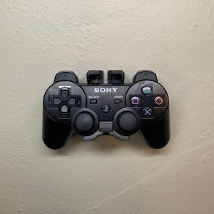 Op de afbeelding: Een zwarte Sony PlayStation 3-controller gemonteerd aan een muur met een 3D-geprinte muurbevestiging. De controller heeft de standaardknoppen, inclusief het PlayStation-logo, select, start en de richtingstoetsen.