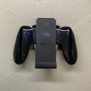 Puede incluir: Un soporte de pared negro para mando de Nintendo Switch con el logotipo y el texto de Nintendo Switch.