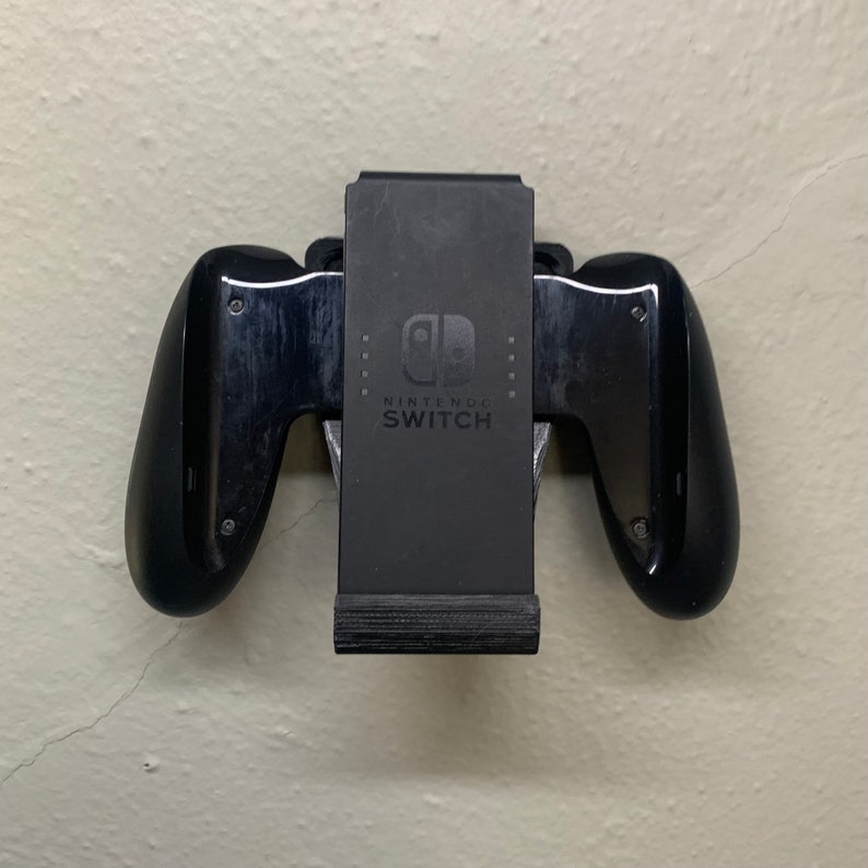 Puede incluir: Un soporte de pared negro para mando de Nintendo Switch con el logotipo y el texto de Nintendo Switch.