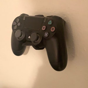 May include: A black PlayStation 4 controller mounted on a white wall. The controller has a black and gray design with buttons for the PlayStation logo, share, options, and the directional pad. The controller also has buttons for triangle, circle, cross, and square.