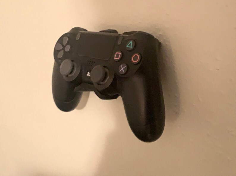 May include: A black PlayStation 4 controller mounted on a white wall. The controller has a black and gray design with buttons for the PlayStation logo, share, options, and the directional pad. The controller also has buttons for triangle, circle, cross, and square.