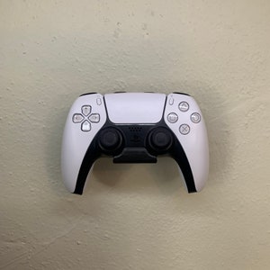 Pode incluir: Um controlador sem fio PlayStation 5 DualSense branco com detalhes e botões pretos. O controlador está montado em uma parede.