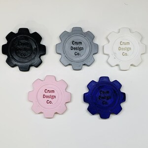 Puede incluir: Cinco posavasos con forma de engranaje impresos en 3D en diferentes colores: negro, gris, blanco, rosa y azul. Cada posavasos tiene el texto "Crum Design Co." grabado en la parte superior.