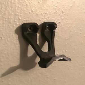 May include: A black plastic wall mount with two screw holes. The mount is designed to hold a gaming controller.