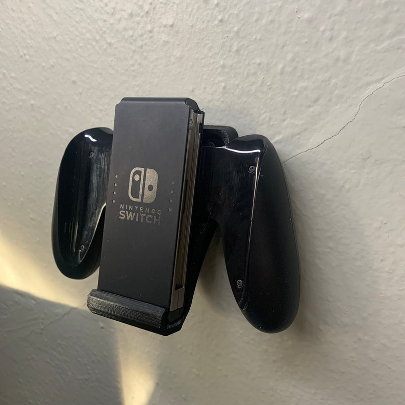 Puede incluir: Un soporte de mando negro para Nintendo Switch con las palabras "Nintendo Switch" impresas en la parte delantera.