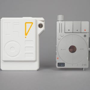 Może przedstawiać: Dwa futurystyczne urządzenia. Jedno jest białe z żółtym trójkątem i okręgiem. Drugie jest srebrne z okrągłym wzorem i przyciskami. Urządzenia mają minimalistyczną, techniczną estetykę.