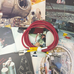 May include: A red guitar cable with yellow ends and white connectors. The cable is coiled and is lying on top of a pile of vintage space-themed photographs.