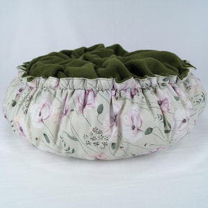 May include: A round pet bed with a green fleece lining and a white fabric exterior with a floral pattern. The bed has a gathered ruffle around the top edge.